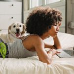 Companion Planting - Relaxed black woman watching laptop near dog on bed